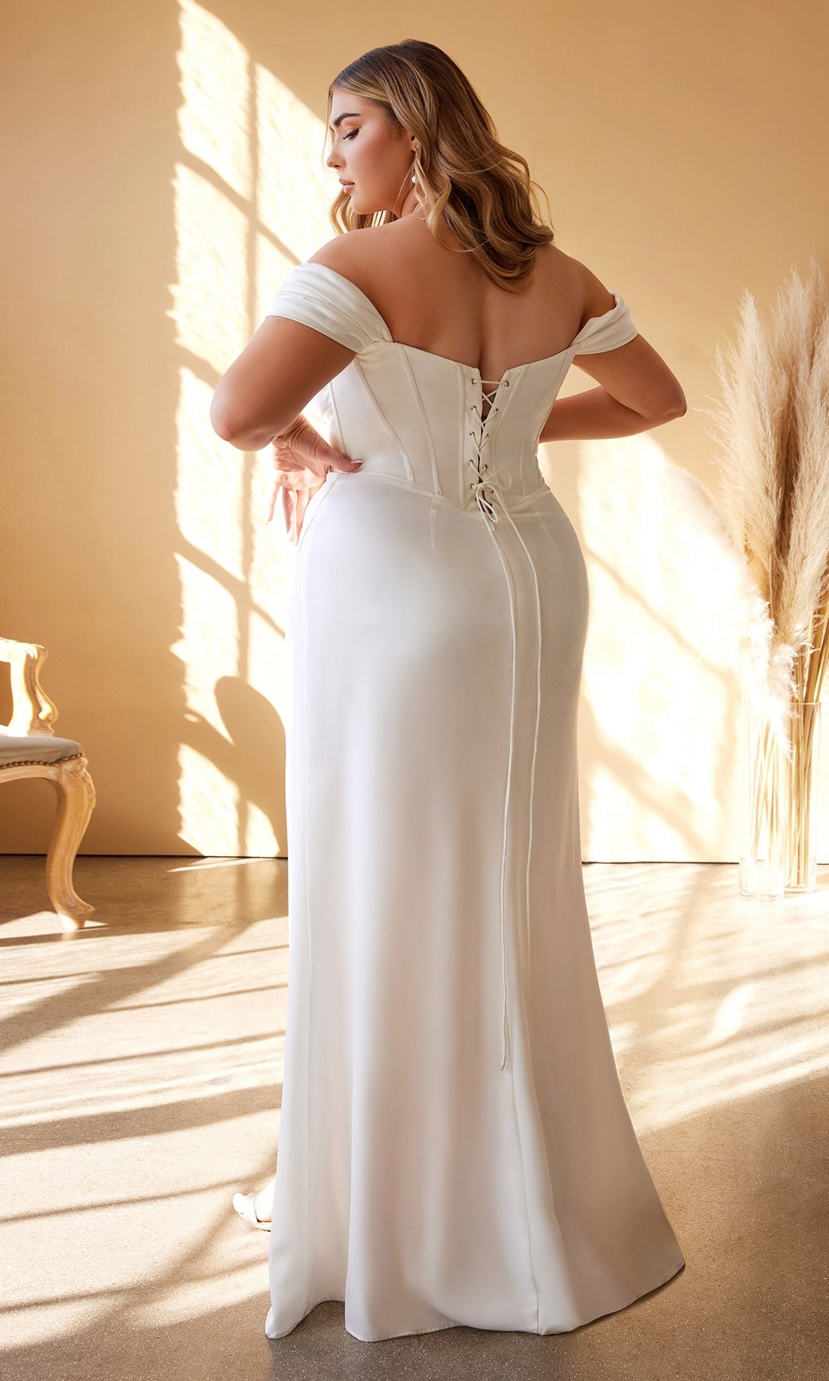 A woman in a sunlit room wears the Off-Shoulder Plus-Size Long White Prom Dress 7484WC, featuring a lace-up back. Facing away, she poses with pampas grass and a chair in the background, highlighting this elegant formal option.